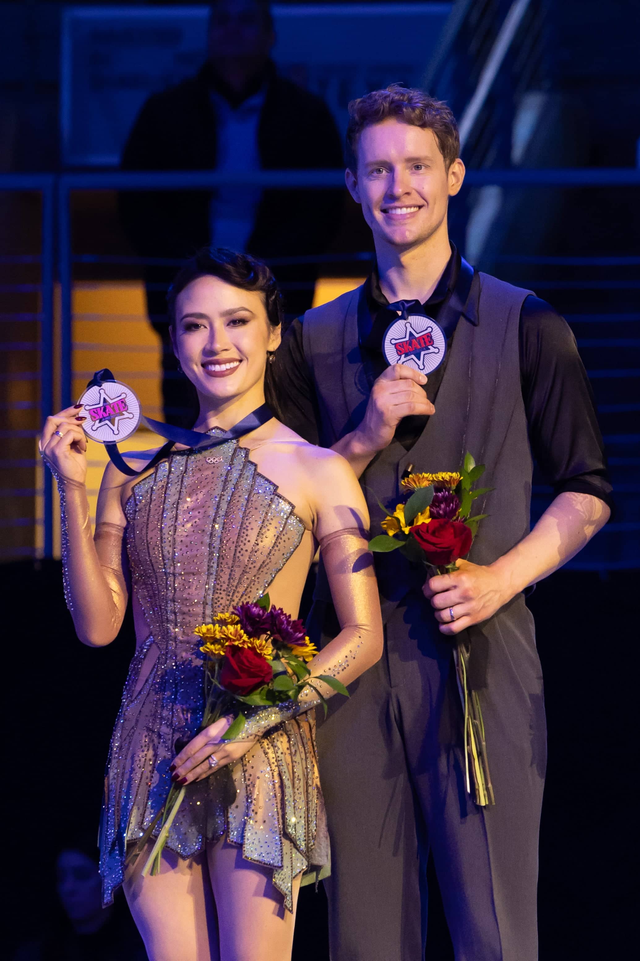 Madison Chock and Evan Bates pose with their Skate America medals on the podium. Madison is a woman with long black hair wearing a costume inspired by the Empire State Building. Evan is a tall man with short blonde hair wearing a black button down shirt with a grey vest and matching pants